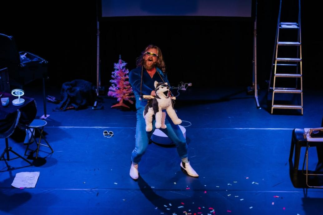 Comedian Paul Currie on stage in a blue jump suit, sunglasses and with a stuffed animal (dog) 
