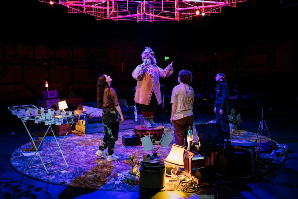 4 actors on stage in the round looking up to a metal sculptural object above them