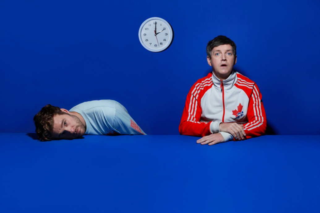 Two men sitting at a table in front of a clock with one slumped over on the table