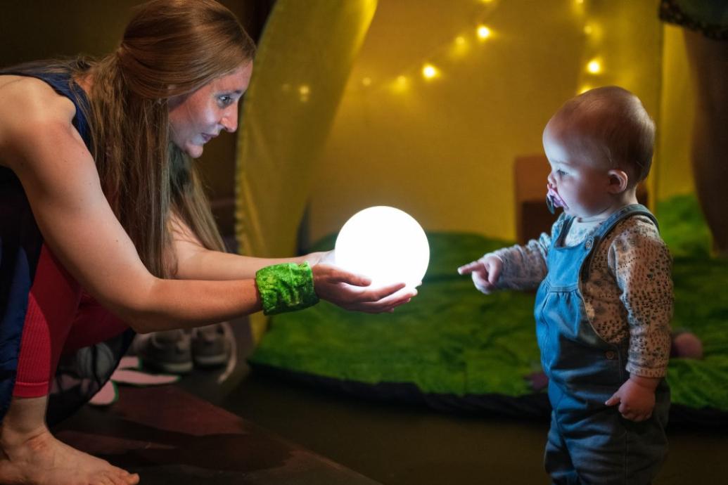 A woman crouching and holding an illuminated globe as a baby touches their finger to it