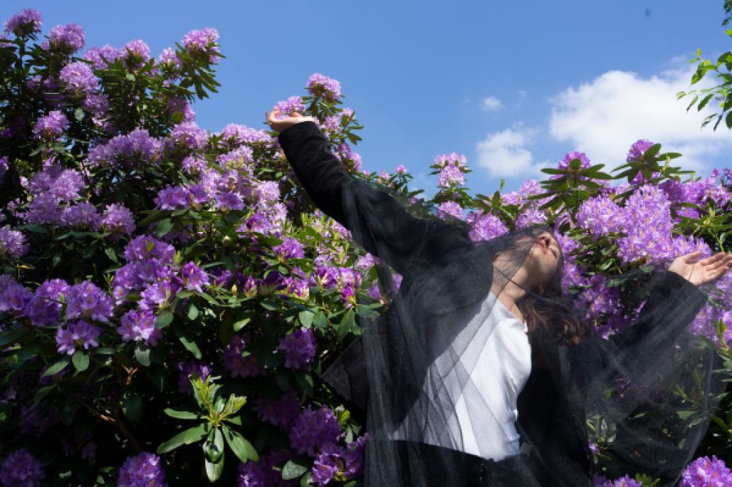 A woman with sheer black material covering her dances in front of a purple flowering bush and blue sky