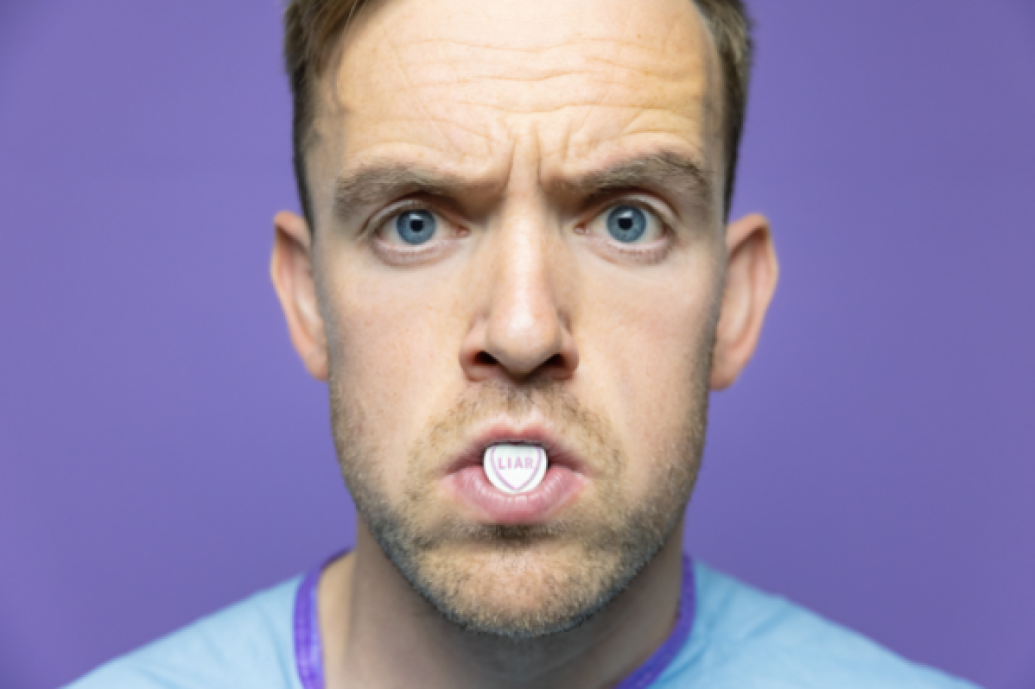 A close up portrait of a man staring straight to camera with a Love Heart sweet in his mouth saying 'Liar'