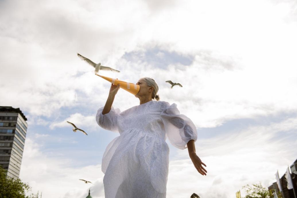 A woman in a white dress holds a paper beak to her face against a cloudy sky in the background