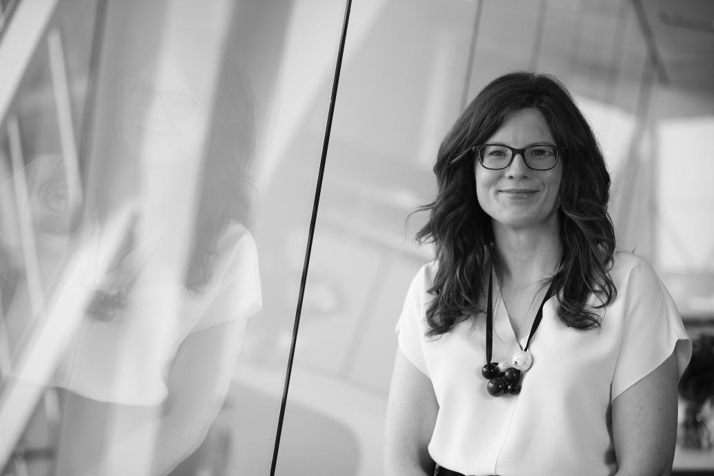 A white woman with long brown hair wearing a white t-shirt and statement necklace with glasses