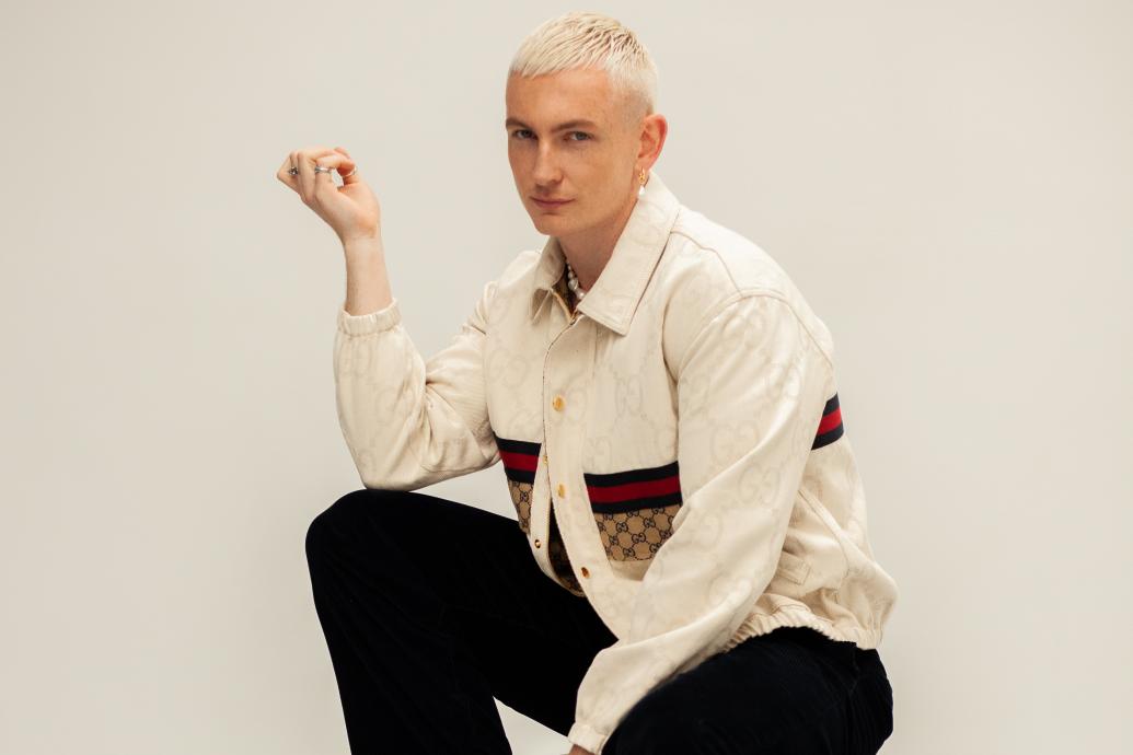 A white name with blond hair sits on a chair looking toward the camera