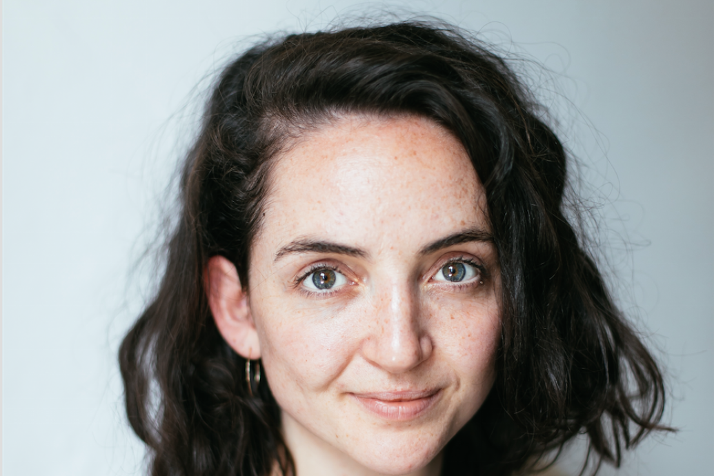 A white woman with shoulder length hair wearing a short sleeved t-shirt and smiling to camera