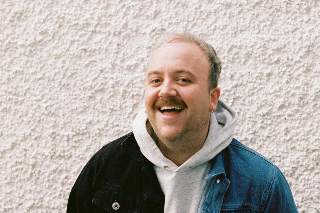 A white man with a moustashe laughing to camera wearing a hoodie and demin jacket in front of a pebble dash wall