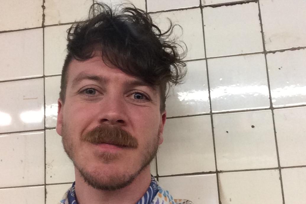 A white man with a moustashe smiling to camera wearing a neck scarf and printed shirt in front of a tiled wall