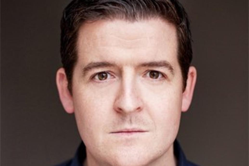 Portait of a white man with dark brown hair wearing a blue shirt looking directly to camera