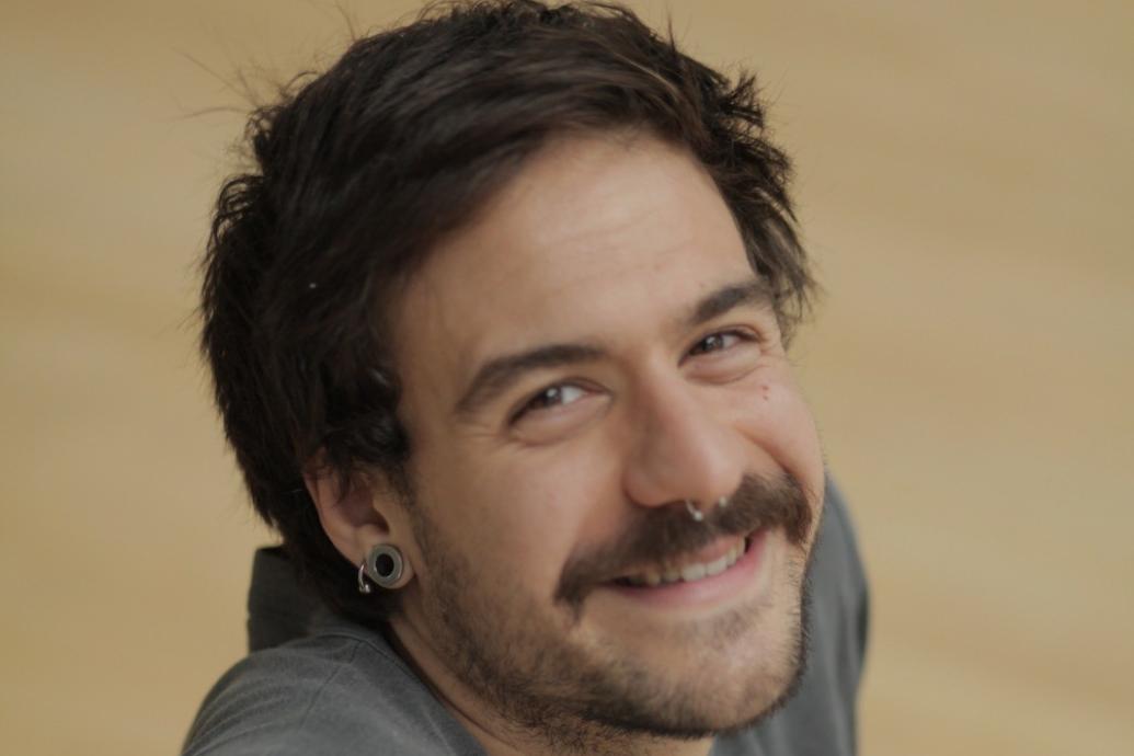 Person with short brown hair and moustache with nose ring smiles to camera