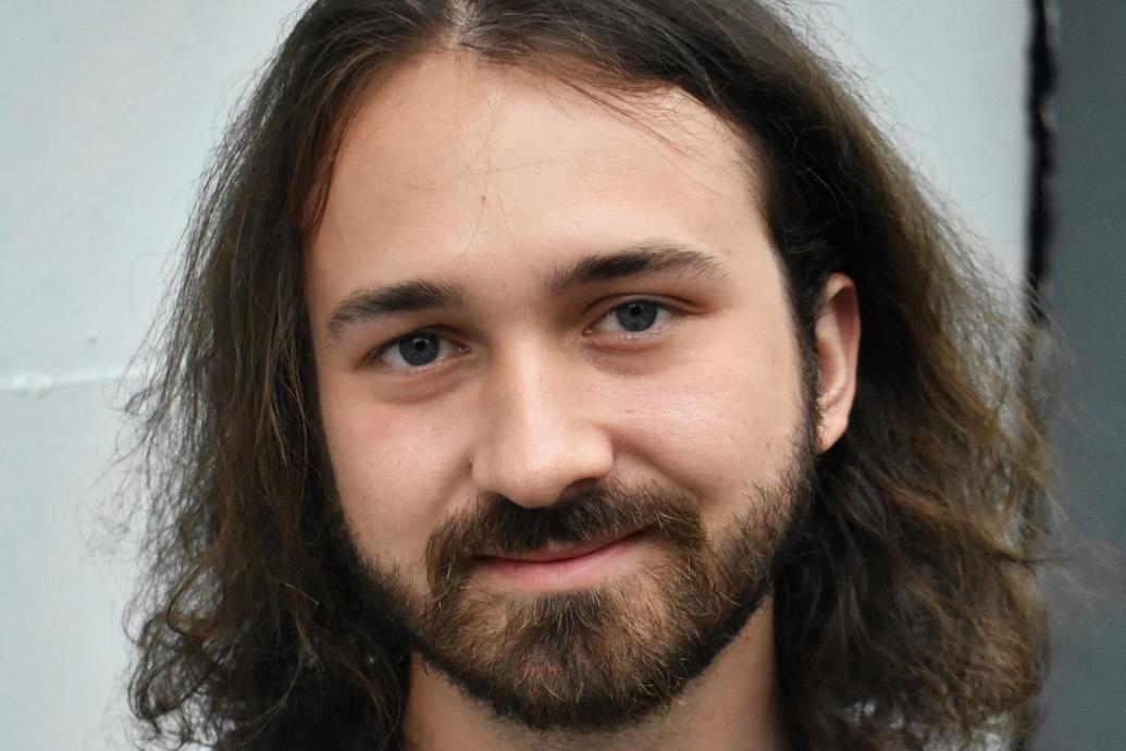 White man with long brown hair and beard wearing printed shirt smiles to camera