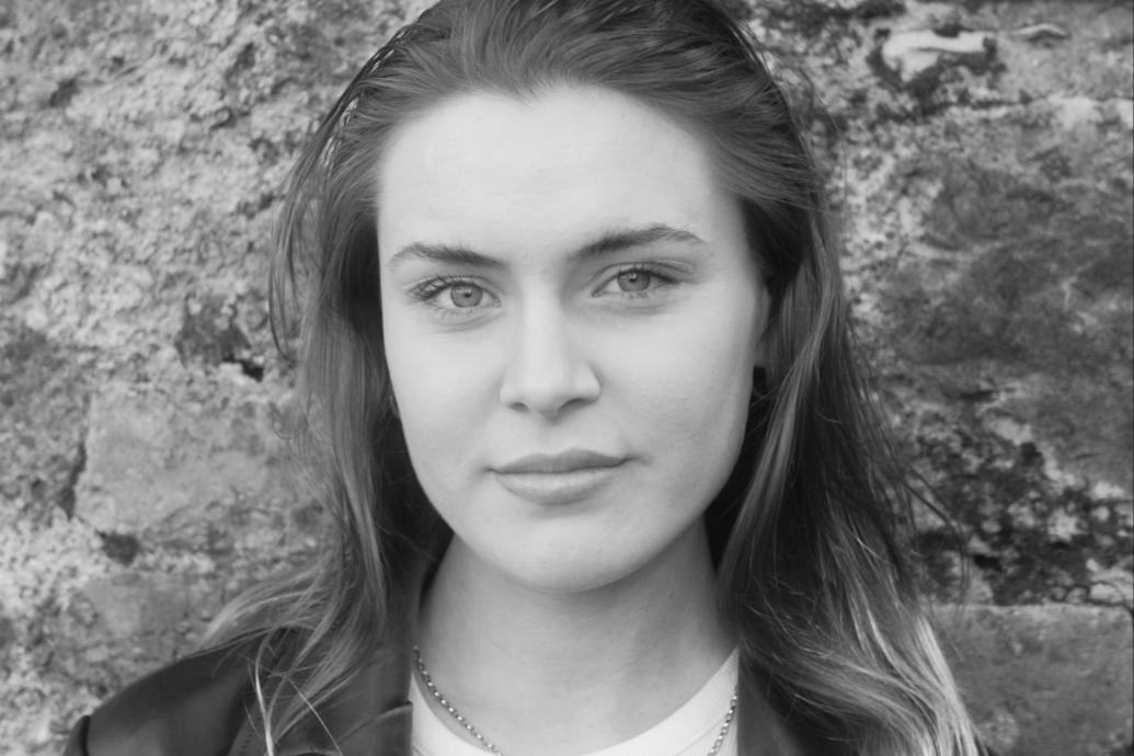 A black and white headshot of a woman standing in front of a stone wall.