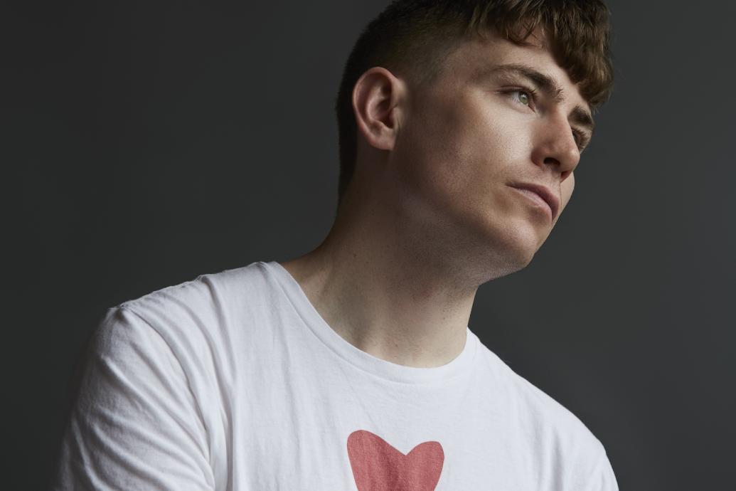 A headshot of a person wearing a white t-shirt with a heart and the word 'The' visible, against a dark background.