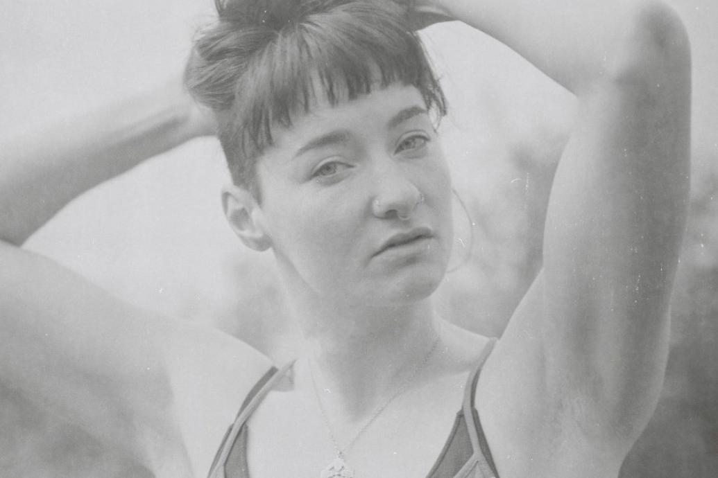 A faded black and white image of a women wearing a vest with arms raised up on her head