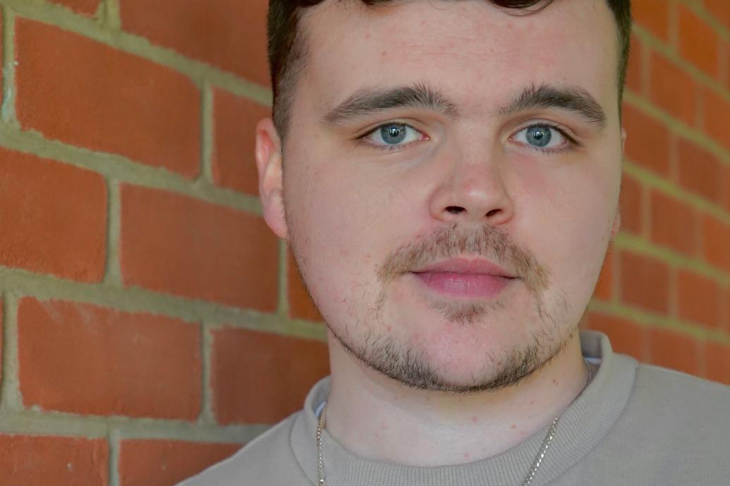 A headshot of a person wearing a gray top standing against a red brick wall.