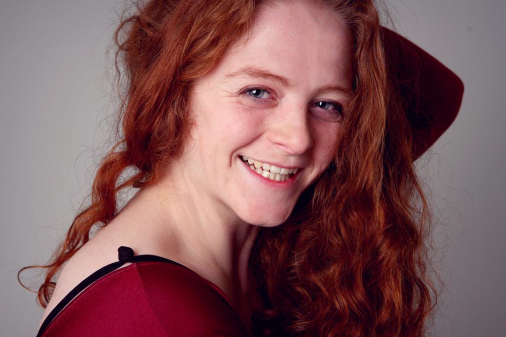 A headshot of a person who is smiling and wearing a read top and with long red hair