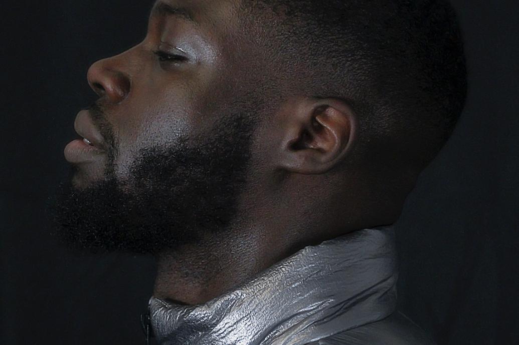 A side view headshot of a man wearing a silver top against a dark background