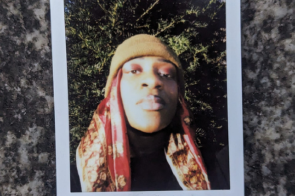 Polaroid photo of a person wearing a beanie and a patterned scarf, lying on their back against foliage, with a granite texture background.