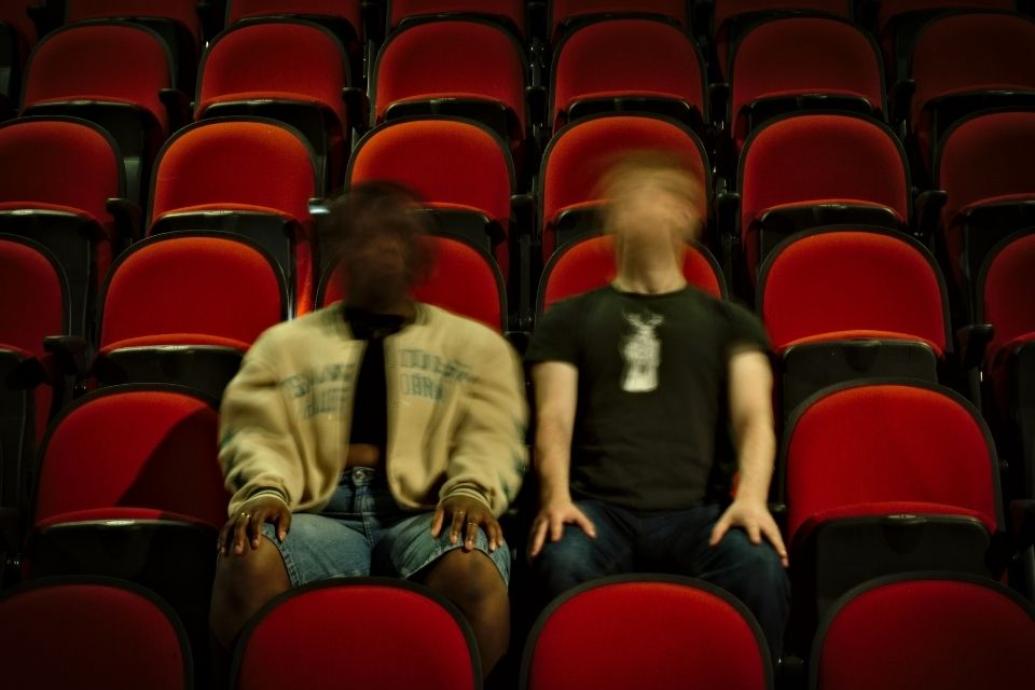 Two people sit side by side in the middle of an empty theatre with red and black seats. Their faces are blurred in motion, creating a ghostly, disorienting effect. One wears a light jacket and shorts, the other a black t-shirt. The lighting is low, adding to the eerie, surreal mood.