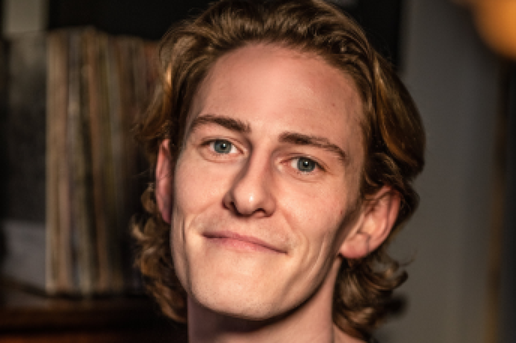 A person with wavy, shoulder-length hair smiles softly at the camera, wearing a dark shirt. The background is dimly lit.