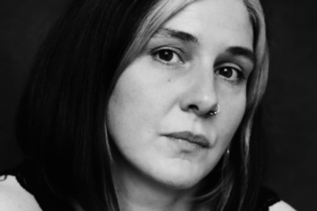 A black and white portrait of a person with shoulder-length dark hair, a white streak, and a nose ring, conveying a contemplative and solemn expression.