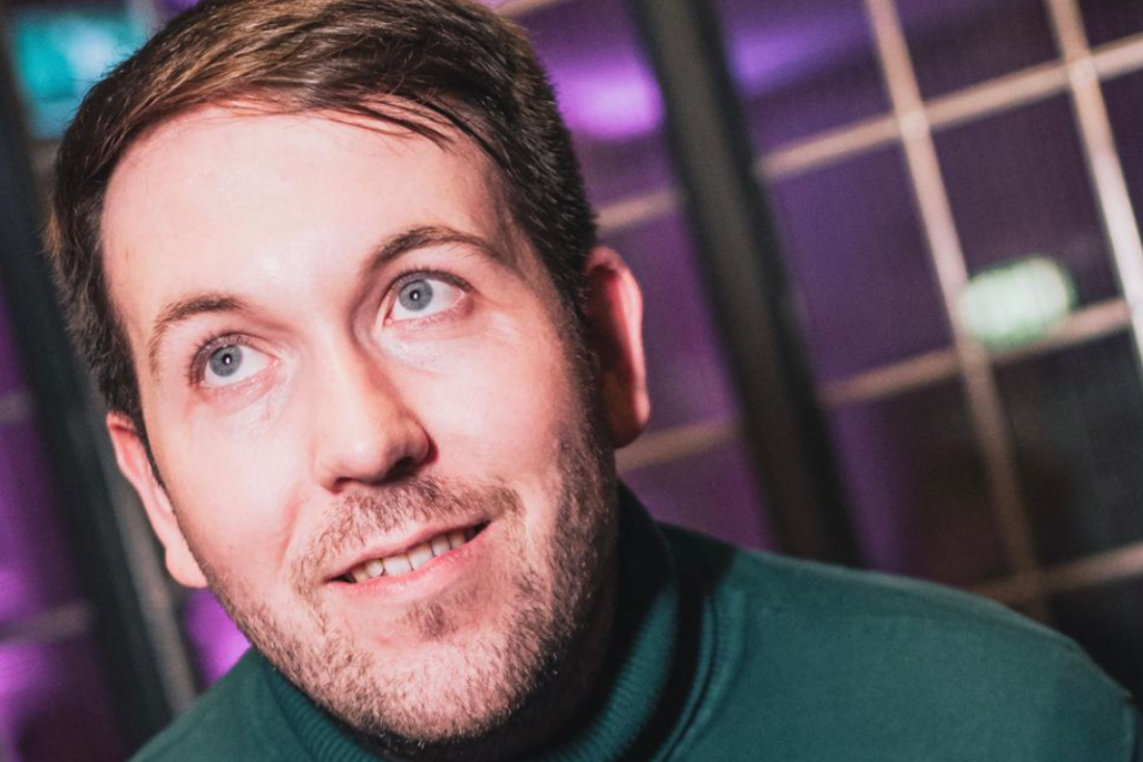 A man with brown hair and a beard, wearing a green turtleneck, looks upward and smiles; purple lighting and grid windows are visible in the background.