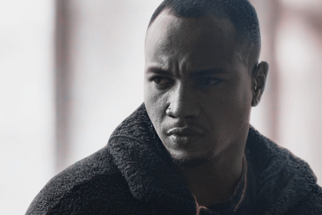 A man gazes intensely to the side in a close-up portrait. He wears a textured dark coat. The background is softly blurred, emphasizing his serious expression.