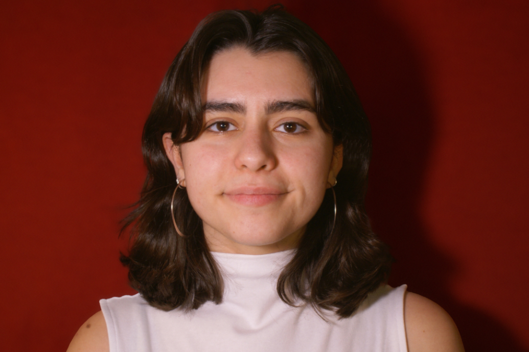 Young woman with shoulder-length dark hair, wearing hoop earrings and a white sleeveless top, stands against a red background, conveying a calm expression.