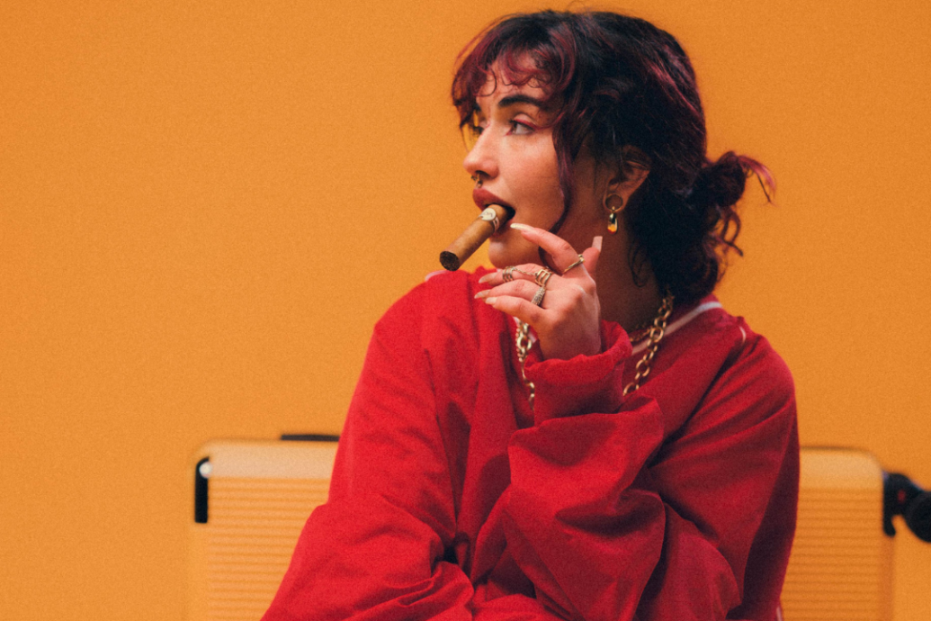 Woman in a red sweater holds a cigar, sitting against a bright orange background. She gazes thoughtfully to the side, conveying a relaxed mood.