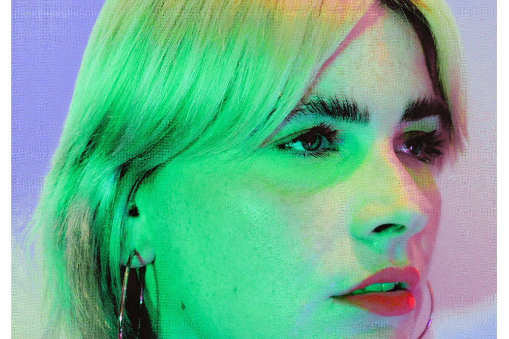 A person with blonde hair is shown in close-up with a contemplative expression, bathed in vibrant green and pink lighting that creates a modern, artistic mood.