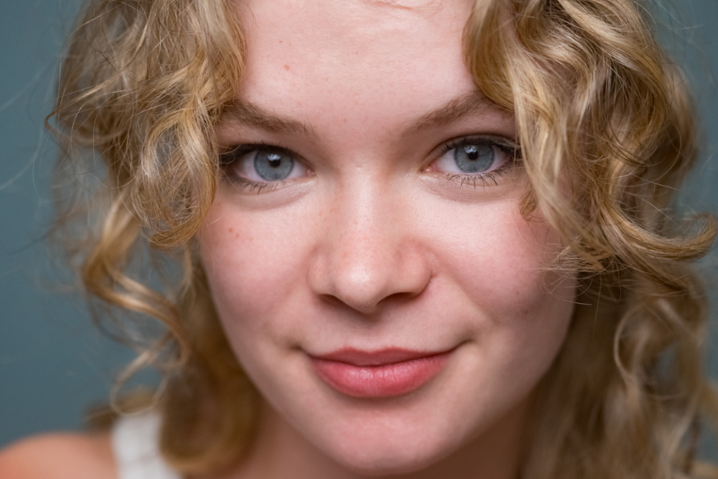 Young woman with curly blonde hair smiles gently at the camera. She has blue eyes and a soft expression, set against a plain, neutral background.