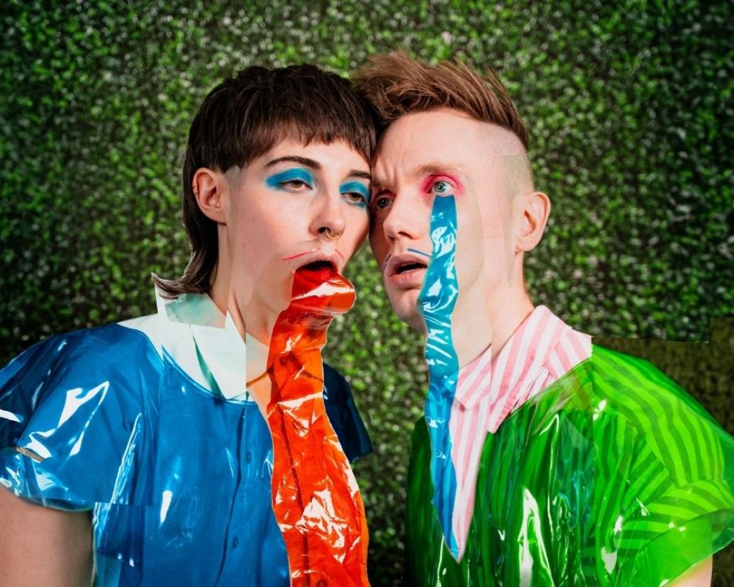 Two people with bold, colorful makeup pose closely against a green hedge backdrop, wearing bright plastic-like clothing with long, crinkled strips of red and blue plastic extending down from their faces like trails.