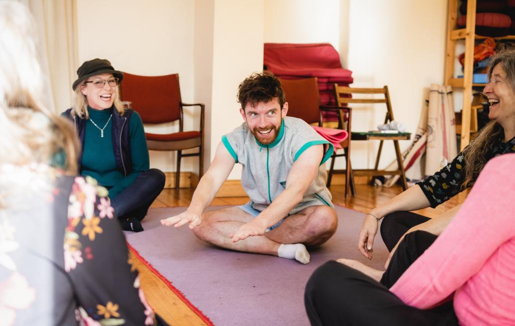 A group of people sitting in a circle on a purple mat, laughing and smiling. In the center, a person sits cross-legged, leaning forward with their hands outstretched