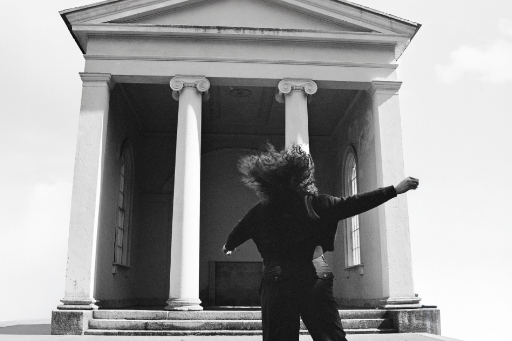 A person with arms outstretched stands before a neoclassical building with tall white columns in a dramatic black-and-white photograph.