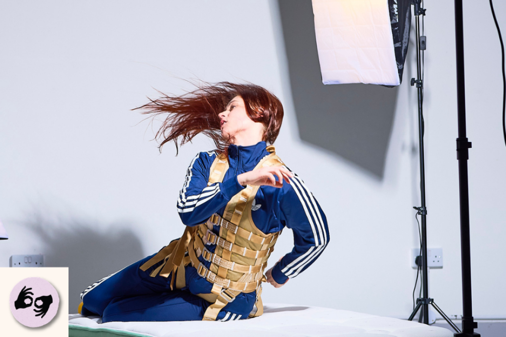 A performer in a blue tracksuit kneels on a white studio floor, turning their head as their hair moves. Visible logo: ISL.