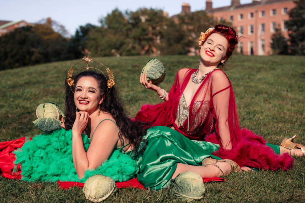 Two performers in bright red and green cabaret-style outfits pose on grass holding cabbages, with a large building in the background.
