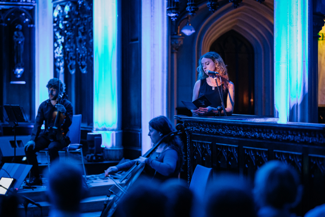 A blue-lit performance in a church-like venue, with musicians seated and a performer speaking at a microphone.