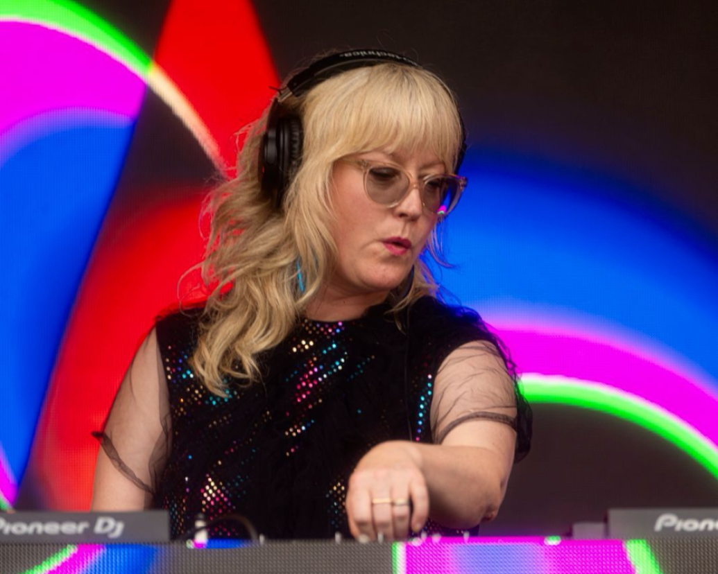 A person with blonde hair, bangs, and glasses wearing headphones and a sparkly black top, operating Pioneer DJ equipment in front of a colorful, abstract neon background.