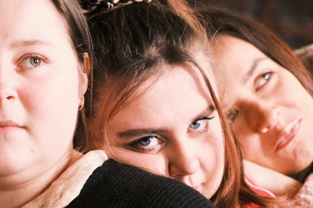A close-up of three people leaning in tightly together, looking toward the camera with expressive facial expressions.