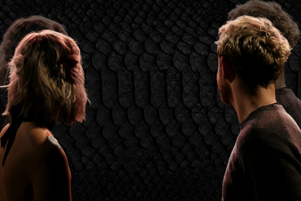 Two people viewed from behind look toward a dark, textured wall, their reflections faintly repeated around them.