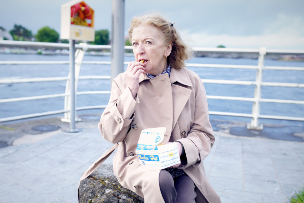 A person wearing a beige trench coat sits outdoors near water, eating food from a takeout box.