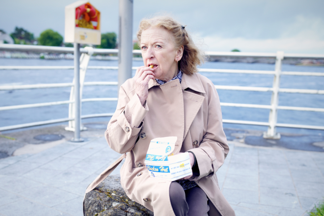 An older person wearing a beige trench coat sits outdoors near water, eating food from a takeout box.