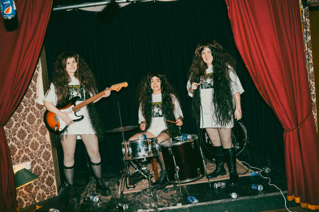 Three performers with long wigs pose as a band on a small stage with red curtains, instruments and crushed cans scattered around them.