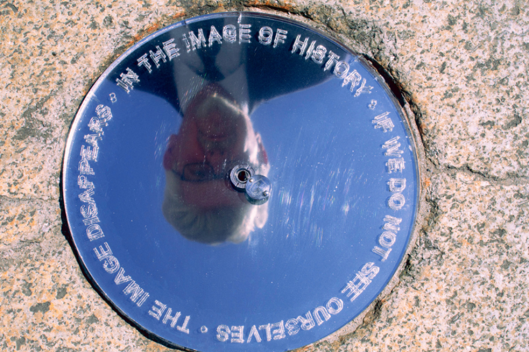 A reflective metal disc set into a stone surface shows an upside-down reflection of a person. Text around the disc reads: “The image disappears in the image of history if we do not see ourselves.” No access or audience icons shown.
