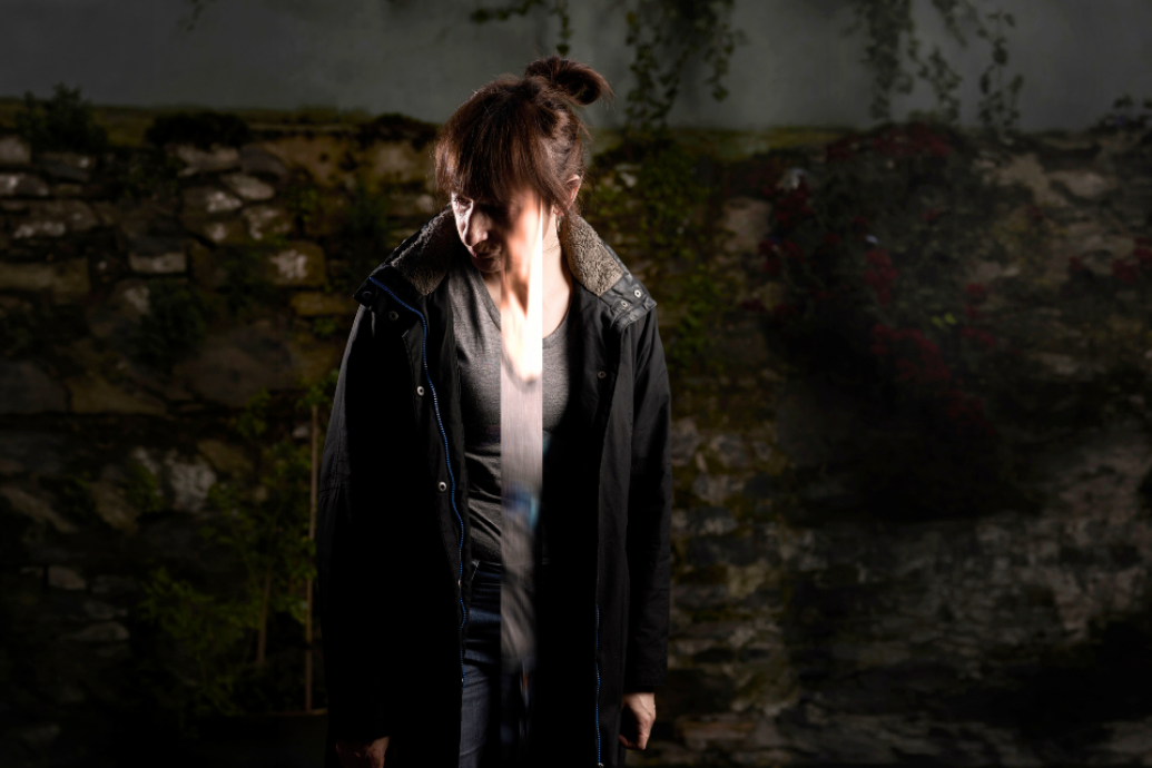 A woman stands in a dark outdoor space with a beam of light passing down the centre of her face and body.