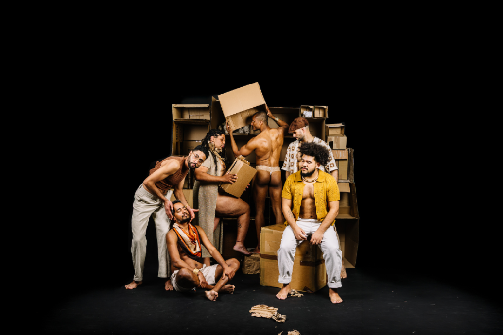 A group of performers gather around stacked cardboard boxes on a dark stage.