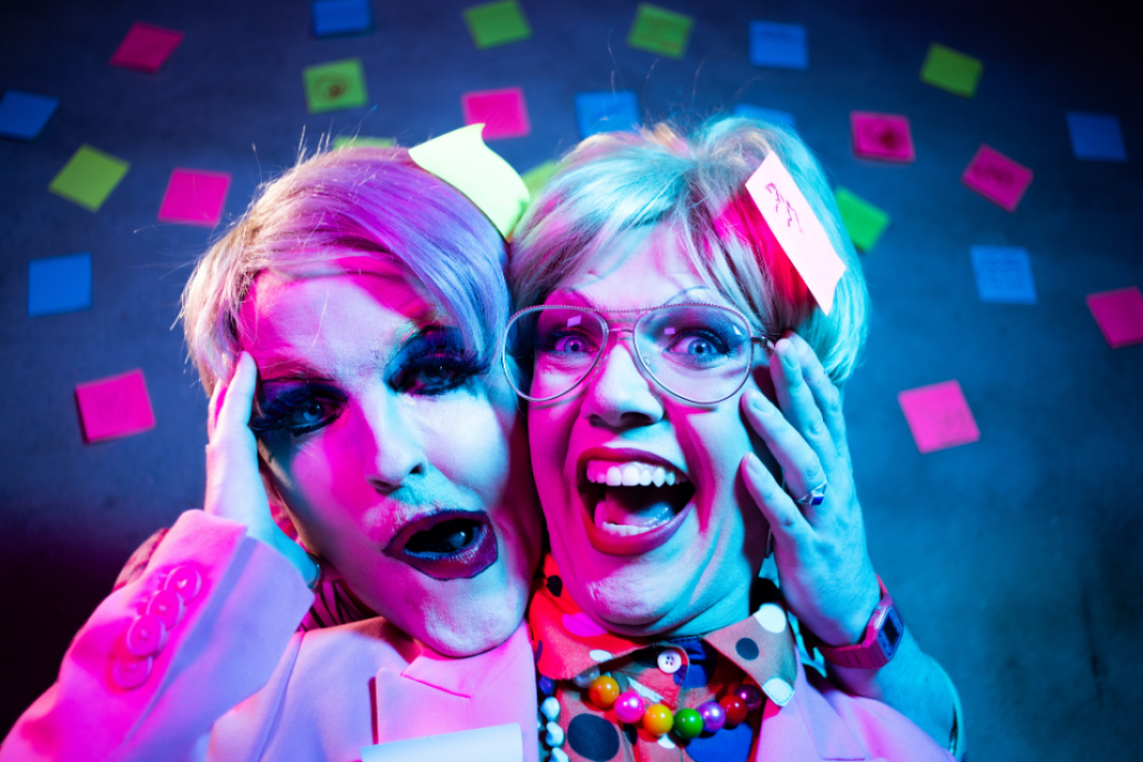Two performers in heavy makeup and colorful attire pose with exaggerated facial expressions under pink and blue lighting against a wall covered in sticky notes.
