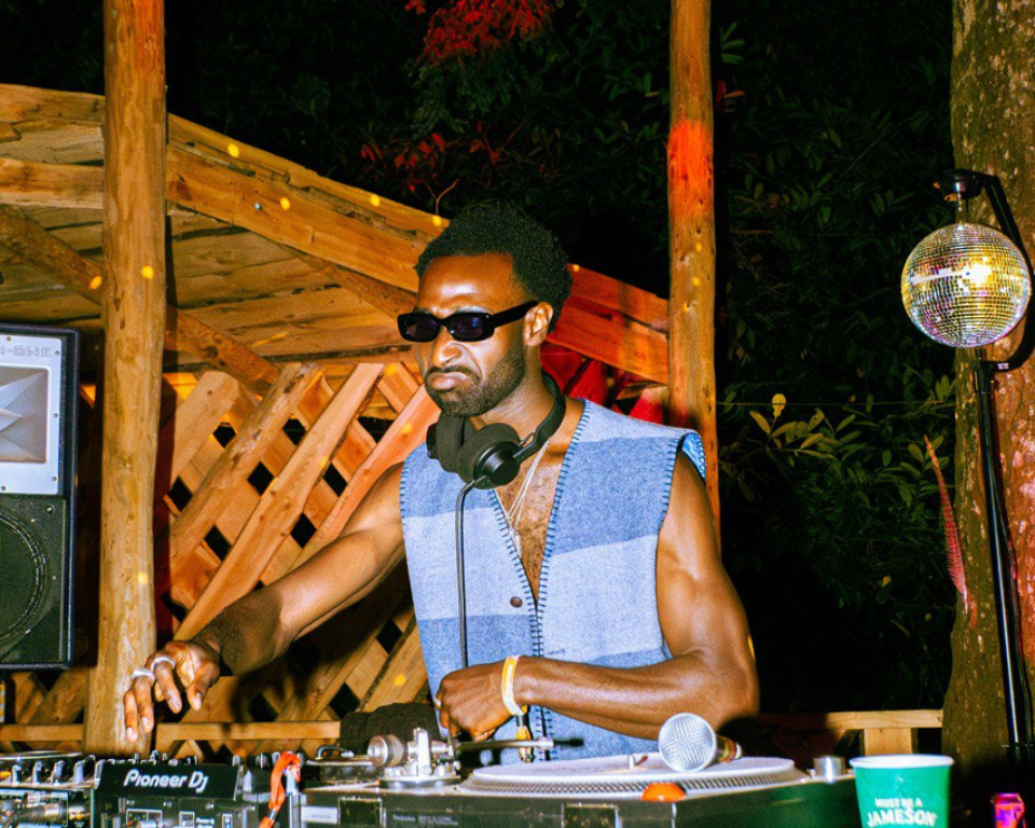 A person wearing sunglasses and a blue patterned vest DJing at an outdoor night venue, with a hanging disco ball on the right and a wooden structure in the background.