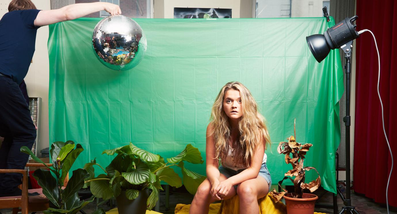 Woman sitting by green screen surrounded by props of plants and a disco ball held in the air by a man